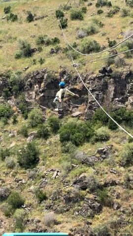 A person wearing a blue helmet and a white zip-up jacket is zip-lining across a rocky canyon. They are suspended by a cable and harness and are in mid-air. The canyon walls are covered with green vegetation and rocks. The sky is blue with some clouds. The person is leaning to their right, arms outstretched. A blue railing is visible in the foreground.