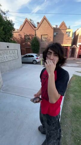 The video shows two young people, one after the other, in what appears to be a residential area with modern brick townhouses. The first person is a young man with long dark hair, wearing a red and black t-shirt, dark patterned pants, and holding a phone, giving a rock-on hand gesture. The second person is a young woman with long blonde hair, wearing a light grey long-sleeved shirt, dark blue sweatpants with white designs, a light blue beanie, light blue shoes, and a black and blue scarf. She is standing in front of a concrete wall with text that reads "A DE LAS OPERATIVAS." The camera angle is low, looking up at both people. The environment is sunny with blue skies.