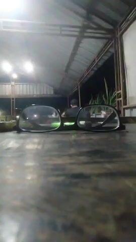 The video shows a close-up of a pair of eyeglasses resting on a wooden table at night. In the background, under a covered structure with fluorescent lighting, a person wearing a dark shirt is seated at a table, facing away from the camera. There are plants visible in the background. The camera is positioned very low, focusing on the eyeglasses.