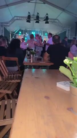 The video captures a lively scene inside a tented venue, likely a festival or party, illuminated by stage lighting. In the background, a band performs on a brightly lit stage, with several people dancing in front of it. The camera is positioned behind a wooden table in the foreground, with chairs visible to the left. The table is cluttered with drinks and what appears to be a small plant in a metal pot, adding a touch of casualness to the setting. The overall ambiance is one of celebration and enjoyment, with a sense of community and shared experience.