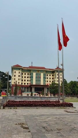 The video shows a wide, tiled plaza in front of a large, multi-story building with a cream-colored facade and a red roof. Two red flags are waving on flagpoles in front of the building. The plaza is partially covered in small patches of green grass. Beyond the plaza are trees, buildings, roads, and vehicles. A mountain range is visible in the background. The sky is overcast. There are a few people visible in the distance.
