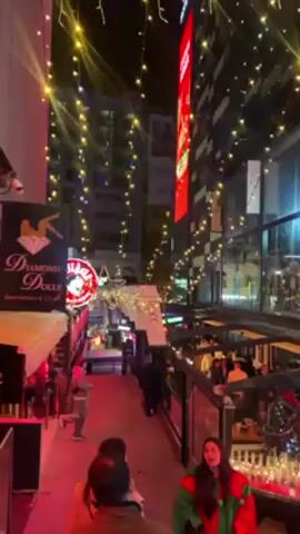 The video shows a bustling outdoor party scene at night. There are many people walking, talking, and hanging out in what appears to be a commercial district. The area is festooned with string lights and neon signs, including those for bars like "Diamond Dolls Gentlemen's Club" and "Nordic Bar". Many establishments are adorned with Christmas-themed decorations. The atmosphere is lively and festive.