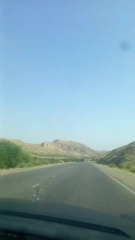 The video shows a view from the dashboard of a vehicle driving on a paved road through a desert landscape with low, sandy hills on either side. The sky is clear and blue. In the foreground, the Mercedes-Benz logo is visible on the hood of the car. The road curves gently into the distance, leading towards a large, flat-topped mountain. Green bushes and vegetation are scattered along the sides of the road and on the hills. The sun appears to be shining brightly. There are no people or other vehicles visible in the video. The time stamp indicates the video is approximately 5 seconds long.