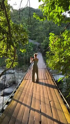 A woman stands on a wooden suspension bridge in a lush, green forest, holding a smartphone. She is facing away from the camera and appears to be taking a picture. The bridge spans a rocky river, and sunlight streams through the trees, casting dappled shadows on the bridge. The woman is wearing a tank top and wide-leg pants. She moves slightly as she takes her photo.