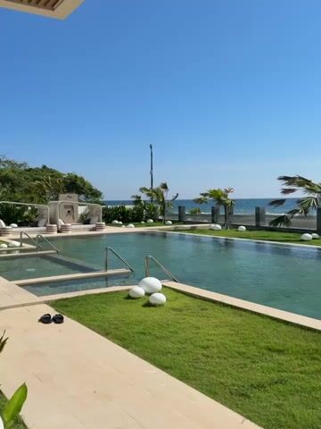The video shows a resort swimming pool area with a view of the ocean. The pool is large and has clear turquoise water. There is a grassy area with white decorative stones and some palm trees that are bending due to the wind. In the distance, the ocean is visible with a clear blue sky above. There are no people visible in the video.