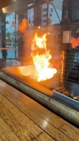 A person in a black shirt and glasses, wearing a black hat with "MIYABI" printed on it, is working behind a glass partition in what appears to be a restaurant kitchen. They are using long sticks to cook something on a flat, hot surface, as evidenced by the flames that flare up periodically. The kitchen is visible through the glass, and outside the restaurant, buildings and streetlights can be seen. Next to the chef is a stack of black bowls. The counter the video is recorded from is made of light-colored wood.
