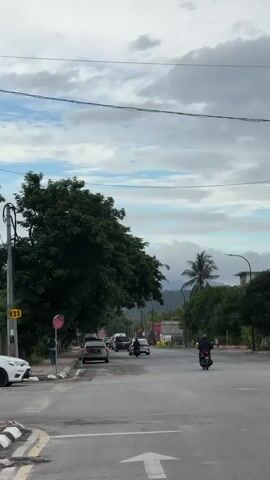 The video shows a moderately busy street intersection under a cloudy sky. There are several cars and motorcycles on the road, with some vehicles moving and others parked. A large tree is visible on the left side of the frame, and more trees line the right side of the road. In the background, there are buildings and hills. The road has clear lane markings, including an arrow indicating a direction of travel. The overall impression is of a typical urban or suburban street scene.