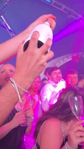 A group of people are celebrating in a nightclub with purple lighting. Someone sprays champagne, and people hold up glasses. One person smokes a cigar. People are smiling and appear to be having fun.