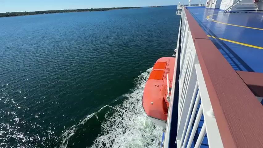 A first-person perspective video shows a ferry sailing through calm blue water under a clear sky. The camera is positioned on the deck of the ferry, looking forward and to the left. On the left side of the frame, the deep blue ocean stretches to the horizon, meeting a distant, tree-lined island. The ferry's side railing, painted a reddish-brown and white, runs along the right side of the frame. A bright orange lifeboat is visible attached to the ferry's hull, with a white stripe on its side and a dark orange hatch. The ferry is leaving a white wake in the water behind it. The deck of the ferry is a vibrant blue with yellow lines painted on it, possibly indicating a helipad or designated walkways. In the distance, the upper decks and structures of the ferry can be seen, including white railings and windows. The overall scene conveys a sense of peaceful travel on the water.