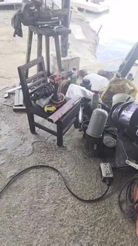 This video captures a somewhat cluttered outdoor workshop area situated near a body of water, likely a river or lake, with lush greenery visible in the background. The scene appears to be in Tambon Mai Khao, Thailand.

The main subjects are a collection of tools and equipment. Prominently featured is a workbench with a vice, various tools, and some rags. To the right of the workbench, a black wooden chair is laden with items including a power drill, a shock absorber, a measuring tape, and a pink metal bar. Beyond the chair, there are several containers, including yellow jerry cans, buckets, and bottles, suggesting the presence of liquids like oil or fuel. A motorcycle wheel is also visible. Further to the right, a welding machine, a coiled green hose or cable, and a pink plastic stool are present.

There is no specific activity or event clearly taking place; the scene appears to be a static display of a workspace.

Notable details include the presence of several power tools, indicating potential mechanical or construction work. The natural light suggests it is daytime, possibly late morning or afternoon given the brightness and slight haze. The location cues, such as the water, dense vegetation, and the style of the structures, are consistent with a tropical or subtropical environment like Thailand. The overall impression is one of a functional, albeit disorganized, outdoor workshop.

No legible text is visible on any of the objects in the frame.