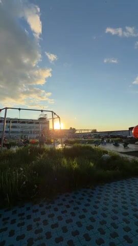 The video shows an outdoor playground area at sunset. The playground features various play structures including swings, geometric climbing structures, and mounds. One mound is painted to resemble a globe.  People can be seen walking and standing on the mounds. The sun is setting behind a building in the background, creating a silhouette effect. The sky is blue with scattered clouds. The ground is paved with blue and gray patterned tiles.  There are patches of grass and weeds growing in some areas.
