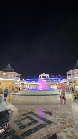 The video shows a nighttime scene in an outdoor plaza with a fountain in the center. The fountain has colorful lights, primarily pink and purple, illuminating the water jets. There are buildings surrounding the plaza, decorated with string lights. Several people are walking around the plaza, including a man in a blue shirt and black shorts on the left, and a couple in green and black outfits walking away from the camera on the right. The ground is paved with patterned tiles. The sky is dark and appears to be nighttime.