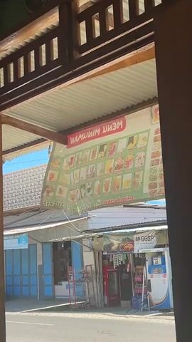 The video shows a view from under a wooden structure, looking out at a street with several small shops. A large banner advertising a drink menu hangs from the roof of one of the shops, its text appearing in reverse. The banner is green with red lettering and displays various drinks and food items. Below the banner, the shops have corrugated metal roofs and open fronts, revealing refrigerators and shelves stocked with goods. In front of one shop, a red Coca-Cola refrigerator is visible, and next to it, a white and blue vending machine. A street runs in front of the shops, and a blue car is parked on the left side of the road. The sky is bright blue.