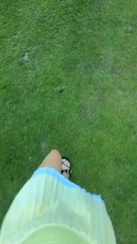The video begins with a first-person perspective of someone walking on a grassy field. Their feet, wearing sandals, are visible in the foreground, with a light yellow skirt. The camera then pans up to show more of the green, open field. The following shots continue with a first-person perspective, showing the grassy terrain, trees in the distance, and a clear blue sky. The video transitions to show a young woman walking away from the camera, also on a grassy area, with trees and a bright blue sky in the background. The final shot is a wider view of the grassy field with trees and the sky, suggesting an outdoor setting like a park or golf course.