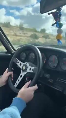 The video shows the view from the driver's seat of a vehicle, with the driver's hands visible on the steering wheel. Through the windshield, a yellow BMW convertible is seen driving on a highway. The camera follows the BMW, capturing its movement alongside the vehicle in which the recording is taking place. The environment includes highway infrastructure, such as guardrails, and a landscape featuring hills and trees under a partly cloudy sky. The video appears to be taken from the perspective of someone racing or following the BMW.