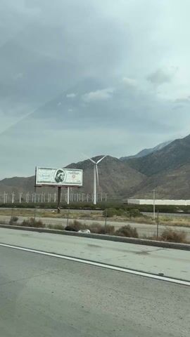 A road trip scene unfolds, captured from within a vehicle as it cruises along a highway. To the right, a vast landscape of desert hills sprawls under a sky partially obscured by clouds. The hills are lined with rows of towering wind turbines, their blades spinning in the breeze. A billboard stands alongside the road, displaying a graphic advertisement. Traffic is light on the highway, with only an occasional vehicle passing by. The perspective is from the driver's side of the vehicle, offering a glimpse of the side mirror and dashboard. The overall mood is one of serene travel amidst a renewable energy landscape.