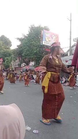 A group of people in traditional clothing are marching down a street. They are wearing brown and orange outfits with yellow sashes. Some of them have red head coverings. The people are walking in a formation and appear to be part of a parade or performance. There are spectators on the sides of the street, some sitting and some standing. In the background, there are buildings and trees. There is also a large billboard with an advertisement. The sky is overcast.