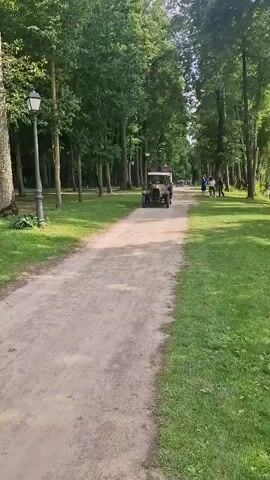 A vintage car drives down a dirt path lined with tall trees and green grass. The car is an open-top model with a light-colored body and dark fenders. It appears to be carrying some luggage on its roof. There are people walking in the distance on the right side of the path. A lamppost stands on the left side of the path. The sun is shining, casting shadows through the trees.