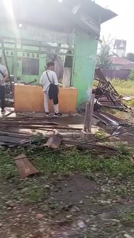 A video shows damage to a structure. The first few seconds show a damaged building with a tin roof that has collapsed, exposing the internal framework. Debris is scattered around the area. The camera then pans to show a sidewalk, a road, and more buildings in the background. The environment appears to be rural or semi-urban with vegetation and trees visible. The remaining seconds show a continuous panning shot of the street and buildings, suggesting a moving vehicle is recording the footage. The scene appears to be an aftermath of some sort of storm or event that caused structural damage. There are no people visible in the video.