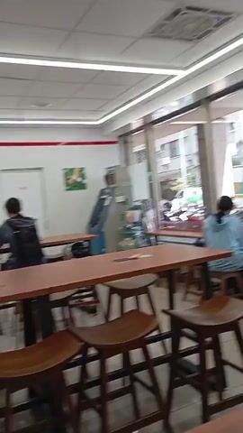 The video showcases the interior of a brightly lit convenience store, likely a 7-Eleven, in Kaohsiung, Taiwan.

The scene opens with a view of the store's dining area, featuring high tables and wooden stools. Large windows look out onto a street where buildings and parked motorcycles/scooters are visible under daylight conditions. Two individuals are seated at separate tables; one person is positioned near the window, and another is further back in the seating area.

A closer shot focuses on a man seated at a high table, engrossed in reading what appears to be a newspaper or document. He is wearing a green collared shirt.

The camera then pans to reveal a wider perspective of the store's interior, looking towards the checkout counter. Shelves stocked with various products line the walls. A prominent "adopt" brand display stand, showcasing what appears to be cosmetics or personal care items, stands in the center foreground. Behind the counter, an employee is briefly visible. Several electronic screens above the counter display advertisements, including an image of a coffee cup with "COCOCAT" text and information written in Chinese script. An ATM is visible in the background against a white wall. The store appears clean, organized, and provides a casual, relaxed atmosphere for customers.
