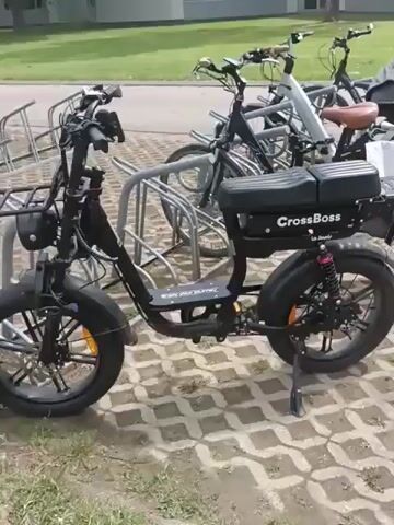 The video shows an electric bicycle parked on a paved surface next to a bike rack. The electric bicycle is black with thick tires and a dual seat. It has a "CrossBoss" logo on the rear battery pack, which is black and rectangular. The bicycle has a front basket and a kickstand. In the background, several other bicycles are parked in the rack, and a building can be seen behind them. The ground is paved with hexagonal concrete blocks, and there is some grass and foliage visible on the left side of the frame. The lighting suggests it is daytime. No people are visible in the video.