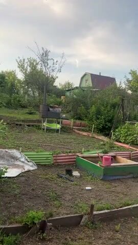 The video shows a slightly overgrown garden with several garden beds. In the foreground, there is a wooden garden bed with green and pink borders. Next to it is a sandbox with a green border and a pink watering can. To the left, there is a tarp and a green corrugated fence. In the middle ground, there is a metal barbecue grill on a stand, and a swing set with lush green plants growing on it. A small, light green house with a dark roof is visible in the background, surrounded by trees. To the right, a dark colored car is parked next to a blue barrel, and a pink watering can is on the ground. The sky is overcast with a few wispy clouds. The overall scene suggests a rural or suburban garden setting.