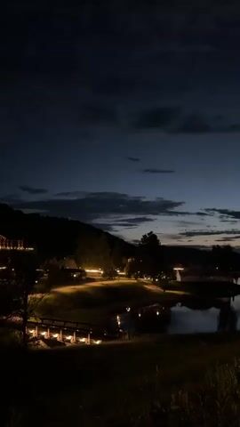 The video captures a tranquil night scene. A body of water, likely a lake or pond, reflects the dim lights that dot the landscape. On the far bank, a silhouette of trees and hills is visible against a dark, cloudy sky. In the foreground, a wooden bridge with illuminated railings spans a section of the water, leading to a path lined with more lights. The overall atmosphere is serene and peaceful, suggesting a quiet evening in a rural or park-like setting.