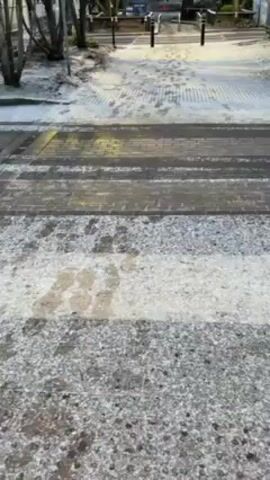The video shows a street covered in thin snow or frost. The camera pans slightly to the right, revealing a crosswalk, a parking area with tire tracks in the snow, a building with blue signage, and a car covered in snow in the distance. The overall scene suggests a cold, possibly early morning, environment.