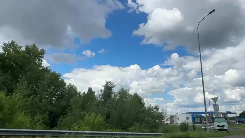 This media file shows a view from a vehicle moving along a road in Chernovskii, Russia. The scene is dominated by a bright blue sky filled with fluffy white clouds, suggesting a pleasant, possibly sunny, day with some cloud cover. Tall streetlights and slender poles are visible, some with overhead wires. On the left, there's a dense line of green trees. To the right, a gas station is in view, featuring a blue and white canopy over gas pumps, a building with signage, and several cars. A white truck with a cylindrical tank is parked near the gas station. There are also some traffic signs, including a red and white circular "no entry" sign. The overall impression is one of a suburban or roadside environment under a dynamic sky. The time of day appears to be daytime, likely mid-morning or afternoon, given the brightness and the quality of light. No people are distinctly visible, but the presence of vehicles and the gas station imply human activity in the area. The visible text includes "Gazpromneft" on the gas station canopy.