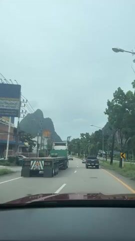 The video shows a first-person perspective from inside a vehicle driving on a two-lane road. The road is bordered by trees and vegetation on both sides. In the distance, a large, distinctive mountain peak is visible. A truck with a flatbed trailer is in the left lane, and several other vehicles, including cars and a pickup truck, are ahead and in the right lane. The sky is overcast, suggesting cloudy or rainy weather. The overall environment appears to be a rural or suburban area with some commercial buildings visible on the left side of the road.