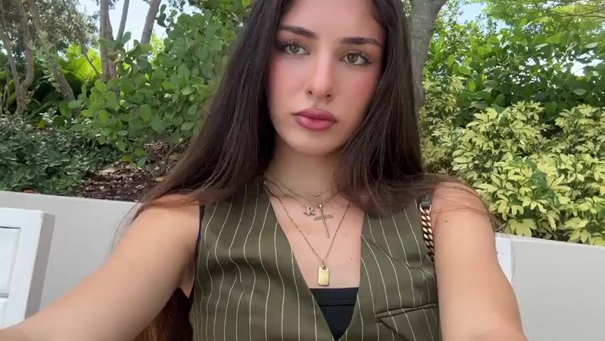 A woman with long, dark hair and noticeable makeup is sitting outdoors. She is wearing a green pinstripe vest over a black top and multiple necklaces, including a cross and a pendant. She is looking towards the camera, which appears to be held by her. In the background, there is lush greenery and a white structure.