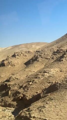 The media file shows a sweeping, panoramic view of a desert landscape with rolling hills and rocky outcrops under a clear, bright blue sky. The terrain is predominantly sandy and arid, with sparse, dry vegetation clinging to the slopes. Shadows cast by the hills indicate it is likely mid-morning or late afternoon, with the sun positioned at an angle. The scene conveys a sense of vastness and natural beauty, characteristic of a desert environment. There are no people or man-made objects visible in the frame. The overall impression is one of a remote and untouched natural setting.