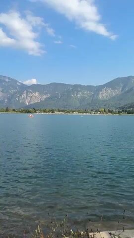 The video shows a scenic view of a large, calm body of water, likely a lake, with a mountainous landscape in the background. The water is a clear blue, and the shoreline in the foreground is rocky and covered with vegetation. The sky is bright blue with scattered white clouds. There are a few small, orange objects visible in the water, possibly small boats or buoys. Trees and green reeds are visible along the water's edge and in the foreground. The overall impression is one of a peaceful, natural environment on a sunny day.