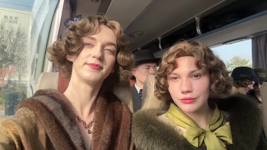 Two individuals with similar hairstyles, makeup, and attire sit side-by-side in a bus, looking directly at the camera. The person on the left has light blue eyes and wears a brown fur coat with a necklace. The person on the right wears a green scarf and a similar fur coat. Several other people are seated in the bus behind them, some wearing period clothing, including one man in a suit and bowler hat. Outside the window is a glimpse of greenery and a sign with Chinese characters and the word "STAGE". The scene suggests a film set or a themed event.