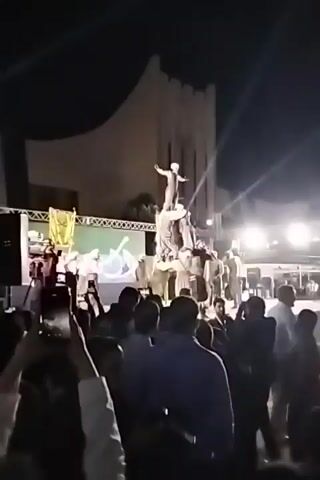 The video shows a group of people gathered at night in front of a building that resembles a castle. A man in a white robe and headdress is standing on top of a pyramid made of people, who are all wearing similar clothing. The crowd is holding up their phones and recording the scene. Behind the pyramid is a stage with a screen displaying what appears to be Arabic writing. There are spotlights shining on the pyramid and the stage.