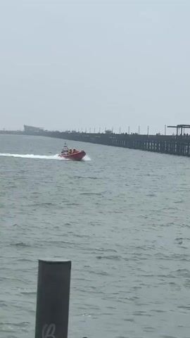 The video shows a red rigid inflatable boat (RIB) moving quickly through the water near a pier. The pier is made of wood and has several people walking along its length. The sky is overcast. There is a dark, possibly metal, post in the foreground, slightly obscuring the lower left corner of the frame. The RIB leaves a white wake as it moves, indicating its speed. The overall environment appears to be a coastal area on a cloudy day.