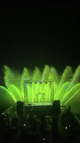 This is a video of a water and light show taking place at night. The main subjects are the illuminated water fountains, which are projected with vibrant green and purple lights. In the foreground, a platform structure with lights is visible, and a crowd of people can be seen gathered below, observing the spectacle. The background shows a distant city skyline with lights twinkling, suggesting an urban or coastal setting. The dark sky indicates it is nighttime, and the water display implies a planned entertainment event. The overall mood appears to be celebratory and awe-inspiring, with the dramatic lighting and water effects creating a visually striking experience. The location is Yeroskipou, Cyprus.