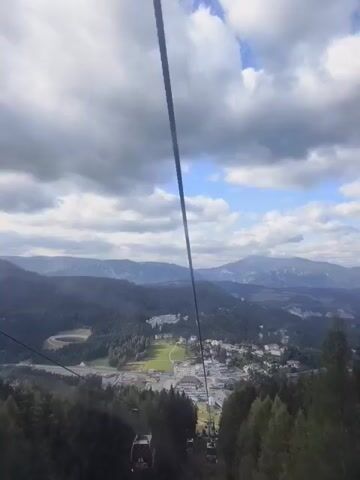 The video is filmed from a gondola lift moving upwards, showing a panoramic view of a mountainous landscape with a village nestled in a valley. The sky is cloudy with patches of blue. The gondola is red and white with the number 17 visible on it. A thick black cable is prominent in the foreground, running vertically through the frame. Below, a winding road and various buildings, including what appears to be a hotel or resort complex, are visible. The valley is surrounded by dense, dark green coniferous forests, and further in the distance, hazy blue mountains rise against the horizon. The movement of the gondola is smooth and steady.