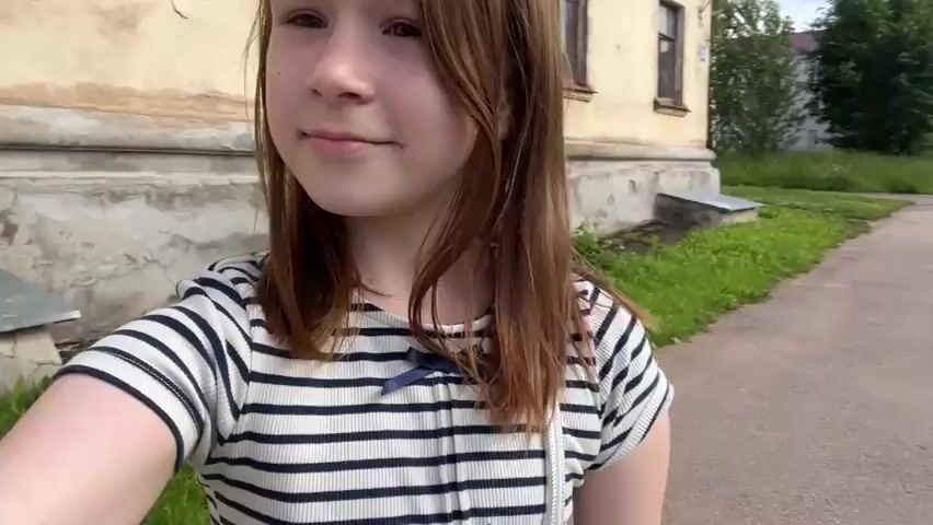 This is a short video of a young girl walking outside, possibly on a sidewalk next to a road. She is the main subject, facing the camera with a slight smile. She has long brown hair and is wearing a black and white striped t-shirt with a small blue bow. In the background, there are residential buildings, trees, and a road with some grass areas.

The activity appears to be a casual walk. The girl briefly touches her hair at one point. There are other people visible in the background, walking behind her on the sidewalk. One woman is wearing a black top and grey pants, carrying a bag with colorful items. Another person, dressed in a grey hoodie and dark pants, is further behind.

The scene suggests it is daytime, with bright lighting and what appears to be a cloudy or overcast sky, though it's not definitively clear. The setting seems to be a suburban or urban residential area in Boksitogorsk, Russia, based on the architectural style of the buildings and the overall environment. The mood of the girl appears calm and perhaps a little playful. No visible text is present in the video.