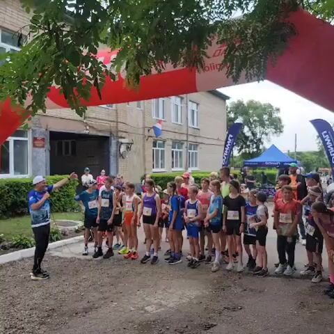 A group of young runners, both boys and girls, are lined up at a starting line. They are wearing athletic clothing and bib numbers. A man in a blue shirt and white hat is speaking to them, possibly giving instructions or encouragement. Behind the runners, there is a building and some trees. An inflatable archway, likely a finish line, is visible in the foreground. The scene appears to be a race or sporting event for children.