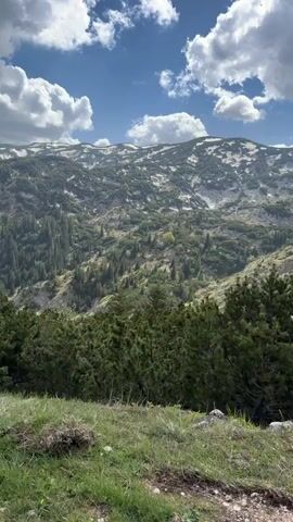 The video shows a mountain range with patches of snow. The foreground has evergreen trees, grass, and a shrub. The sky is blue with white clouds. The camera pans slightly. A car door is briefly visible on the left. Voices are audible.