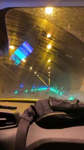 A car travels through a brightly lit tunnel. Rain droplets dot the windshield, and the dashboard is visible. The tunnel's lights are a mix of yellow, green, and blue. A phone is mounted to the windshield. An object, possibly a jacket or bag, rests on the dashboard. The car is moving forward.
