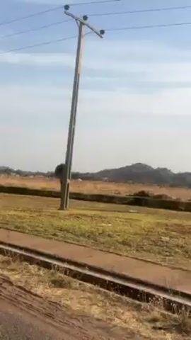 The video shows a rural landscape with dry grass, scattered trees, and hills in the distance, possibly filmed from a moving train. Several unfinished buildings are also visible. The sky is overcast. Power lines are also seen briefly.