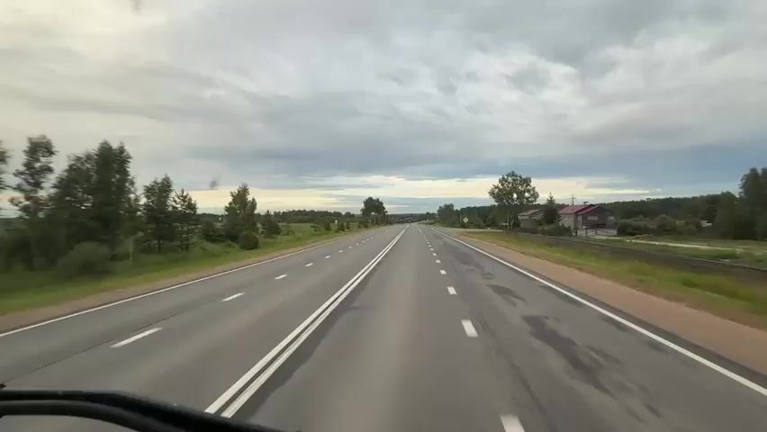 A point-of-view shot from a vehicle driving on a two-lane asphalt road. The road has a double white line down the center and white dashed lines on the sides. The sky is overcast with gray clouds. On either side of the road are green fields and trees. In the distance, there are some houses and buildings, suggesting a rural area or a small village. The camera appears to be mounted low, possibly on the dashboard or in front of the driver. The video captures the forward motion of the vehicle on the road.