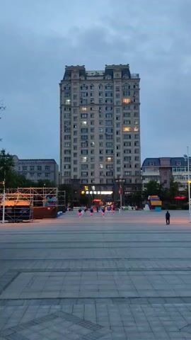 A group of people, mostly women wearing pink skirts and white tops, are performing a synchronized dance in an outdoor plaza. They are spread out in front of a tall, multi-story building with many windows, some of which are lit. To the left of the building, there is a scaffolding structure with lights. The ground of the plaza is paved with large grey tiles. The sky is overcast and appears to be dusk. Several other people are visible in the background, some standing and watching, others walking.
