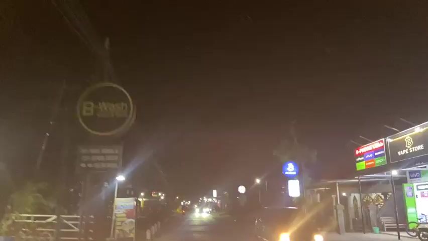 The video shows a first-person perspective from a vehicle traveling down a dimly lit street at night. The street is lined with buildings, some with visible signs and lights. Several streetlights illuminate the road, and the headlights of oncoming vehicles create lens flares. Motorcyclists are present on the road, and some parked motorcycles are visible on the side. The environment appears to be a commercial or residential area with a tropical feel, indicated by the presence of trees. The overall atmosphere is that of a quiet, rural or suburban street at night.