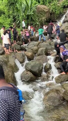 A group of people are gathered around a waterfall, with many of them climbing on the rocks and playing in the water. The environment is lush and green, with trees and vegetation surrounding the waterfall. There are various age groups present, including adults and children. Some individuals are wearing traditional clothing, while others are in casual attire. The waterfall appears to be a popular tourist destination, with a considerable number of visitors present. Some people are taking photos, while others are enjoying the natural scenery and the refreshing water. The overall atmosphere seems to be one of leisure and recreation.