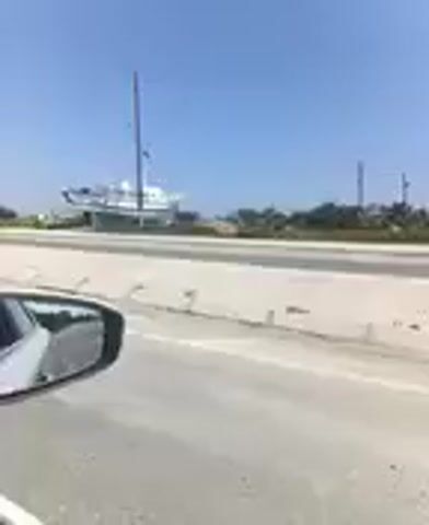 The video shows a view from a vehicle moving along a road. In the distance, a large white boat is visible on land, supported by a structure. The boat appears to be a yacht with multiple decks. Several tall poles, possibly utility poles or mast-like structures, are scattered in the background. The sky is clear and blue, indicating a sunny day. The road surface is visible in the foreground, along with a concrete barrier. The side mirror of the vehicle is partially in view.