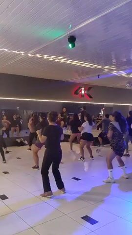 The image depicts an energetic indoor group fitness class taking place in what appears to be a dance or aerobic studio in Tacna, Peru. Approximately 15-20 women, predominantly adults, are actively engaged in a synchronized exercise routine, likely a dance fitness session, with their arms raised and bodies in various dynamic poses. They are dressed in comfortable athletic wear, including shorts, leggings, t-shirts, and sneakers, suggesting a high-energy workout.

The participants are mostly facing away from the camera, indicating they are following an instructor positioned at the front of the room (who is not clearly visible). The studio boasts a clean, white tiled floor with subtle darker square patterns, reflecting the vibrant overhead lighting. The ceiling is white and paneled, fitted with multiple white ceiling fans and black track lights. The background wall is dark grey, featuring a prominent, illuminated "4K" logo in red and white, suggesting the name of the studio or gym.

The space is brightly lit with a mix of white general lighting and colorful accent lights, including noticeable green and purple hues that cast reflections on the polished floor. A small disco ball can be seen in the upper right background, contributing to the lively atmosphere. The overall scene conveys a sense of focused activity and communal exercise.