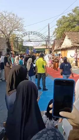 A group of children in costumes, some with elaborate headwear, are walking down a red carpet lined with spectators. A man in a yellow shirt and dark pants appears to be directing the children, gesturing with his hands. Several people, including women wearing hijabs, are watching the procession and some are holding up smartphones to record the event. The scene takes place outdoors on a street with buildings and trees visible in the background. The sky is clear and the lighting suggests it is daytime.